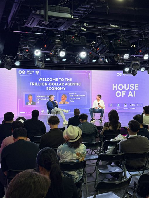 Two men sit on stage at the "House of AI" summit, with a large screen behind them displaying "Welcome to the Trillion-Dollar Agentic Economy" along with the names and titles of speakers Michael Heinrich (CEO & Co-Founder, 0G) and moderator Tim Haldorsboh (CEO of Turtls). The image captures the panel discussion referenced in the post about how AI agents will shape the next economy.