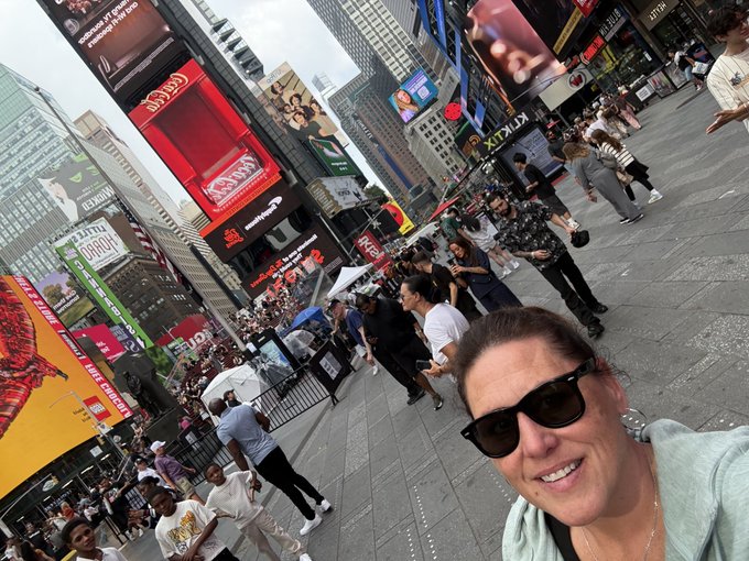 A selfie of a woman wearing sunglasses taken in a crowded Times Square, with large digital billboards and advertisements visible in the background.