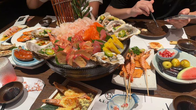 An elaborate seafood feast is spread across a wooden table, featuring a large platter of sashimi, oysters, and prawns on ice, accompanied by grilled skewers, crab, and watermelon.