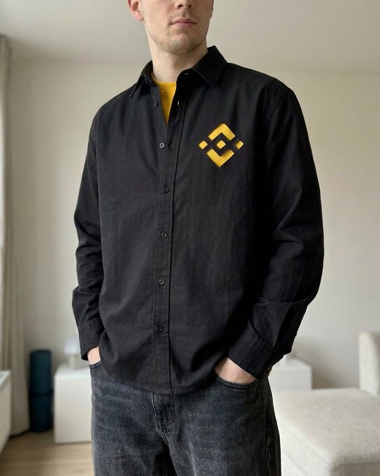 A man wears a black button-up shirt featuring a yellow Binance logo on the chest, paired with a yellow undershirt—turning the crypto exchange's branding into an ironic fashion statement, as if advertising "Binance" like a personal identity or loyalty badge.