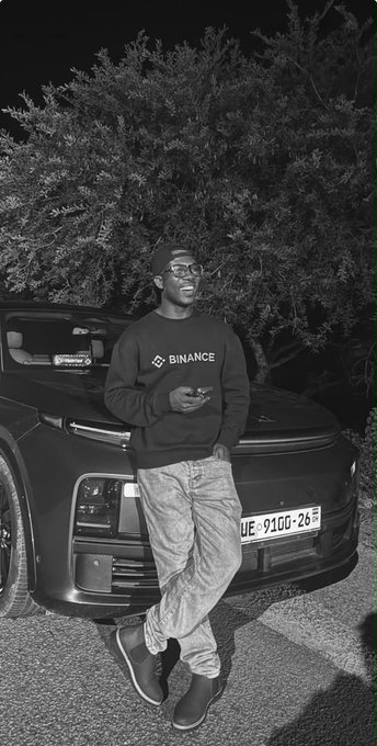 A black and white photo shows a smiling man wearing a 'BINANCE' sweatshirt and cap, leaning against a Rolls-Royce with a license plate reading 'UE-9100-26'.