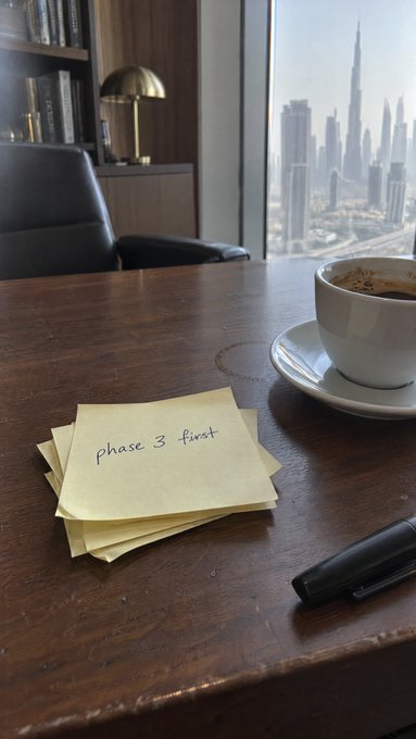 A wooden desk scene featuring sticky notes with handwritten text 'phase 3 first', a cup of black coffee, a black pen, and a view of the Burj Khalifa and Dubai skyline through a window.