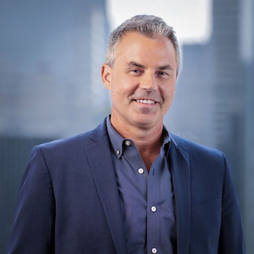 In the image, there's an older man wearing a blue suit jacket with white buttons and a collared shirt. He has gray hair and is smiling at the camera. The background of the photo features a city skyline, suggesting that he might be in a professional setting or attending an event related to finance or technology. The caption provided seems to be unrelated to the main subject matter but includes some interesting information about Bitcoin.