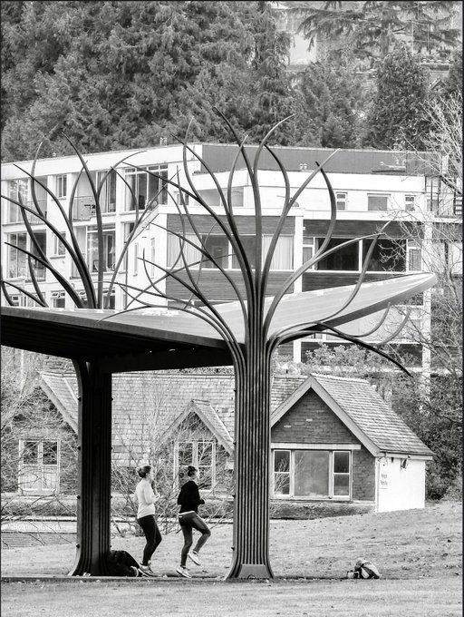 A black and white photograph of two people standing under a large metal sculpture resembling a stylized tree, with a modern apartment building in the background and a small house nearby.