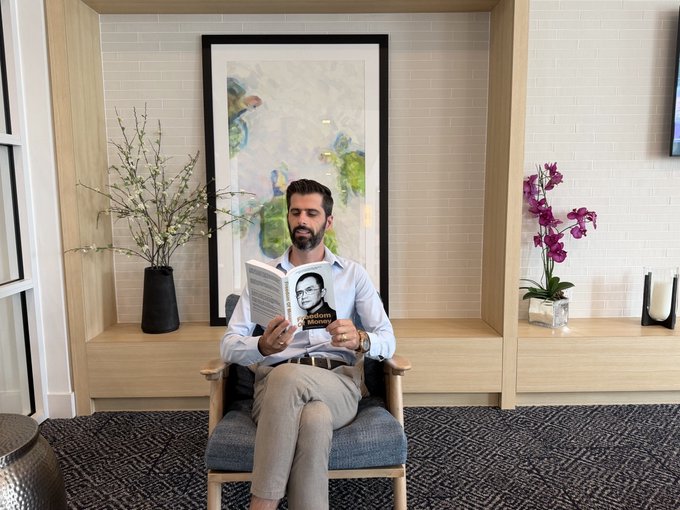The image features a man sitting comfortably in a chair, engrossed in reading a book titled "FREEDOM OF MONEY" by @cz_binance. The scene takes place in an office setting with a TV mounted on the wall behind him and several potted plants placed around the room.
