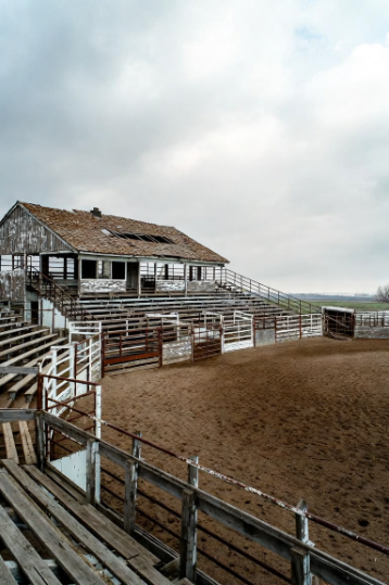 An old wooden barn or livestock building with empty metal pens and bleachers under a cloudy sky.