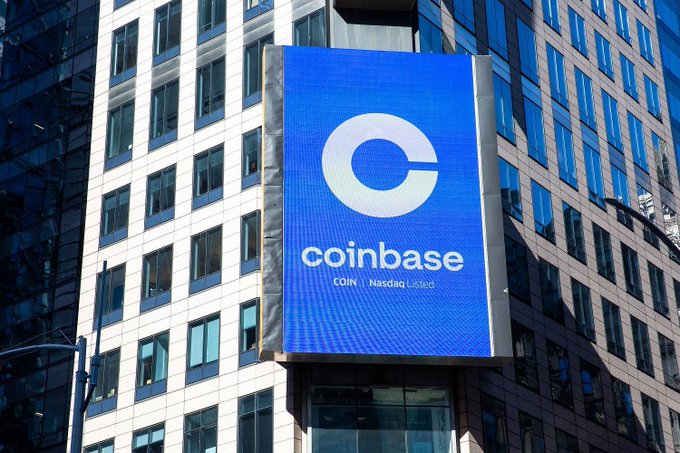 The image features a large billboard for Coinbase that is prominently displayed against a backdrop of tall buildings. The billboard showcases the name "Coinbase" written in white letters, with a blue logo underneath. The text on the billboard reads "Crypto daily update today is quite interesting. OpenAI may reduce token fees. Hong Kong is preparing to regulate stablecoins. Coinbase is reportedly about to expand crypto perpetuals in the US. But at the same time, Glassnode says BTC is heading deeper into the capitulation". This advertisement seems to be promoting a new cryptocurrency or highlighting recent developments within the crypto world.