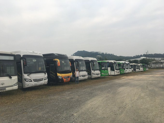 A lineup of various tour buses parked in a lot with a person standing in front of them.