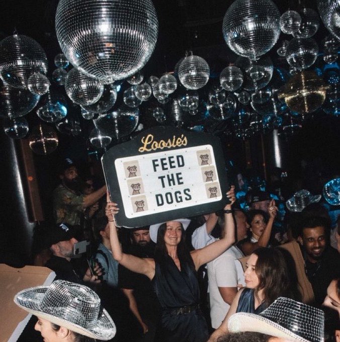 A woman in a crowd at a club holds up a "Loosies Feed the Dogs" sign, surrounded by disco balls hanging from the ceiling and people wearing sparkly hats.