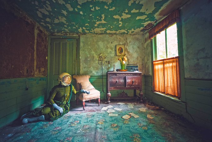 A photograph of an astronaut in a yellow helmet sitting on the floor of an abandoned, decaying room with peeling paint, an old chair, and an antique cabinet.