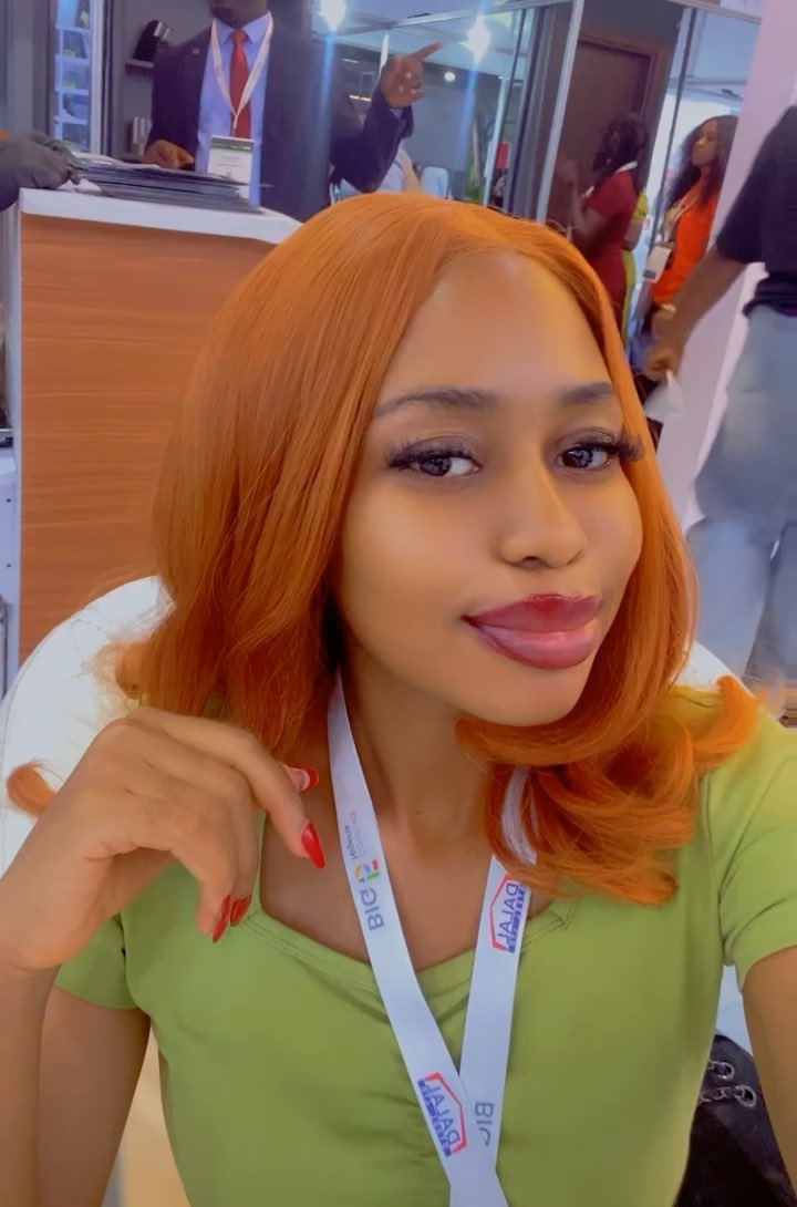 A woman with bright orange hair and long red nails, wearing a green top and a lanyard with 'BIG' and Arabic text, posing at what appears to be an event.