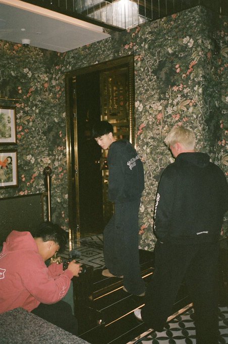 Three young people gather in a hallway with floral wallpaper, where one crouches in a red hoodie to take a photo, while another in a black hoodie stands in a doorway and a third person with bleached blonde hair watches from the side. The casual, behind-the-scenes vibe matches the "Gm Ct We go again today" energy of a fresh day ready to be captured on camera.