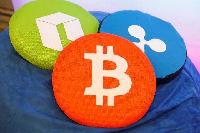 Three circular cushions displaying cryptocurrency logos—Bitcoin (orange), NEO (green), and Ripple/XRP (blue)—rest on a blue fabric surface.