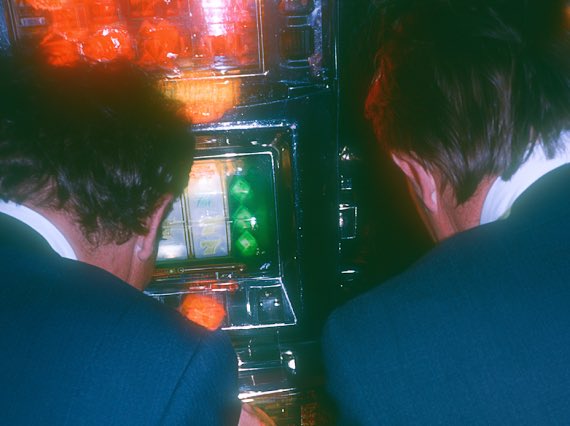 Two people in dark uniforms lean over a slot machine with a glowing green screen, viewed from behind in a dimly lit environment.