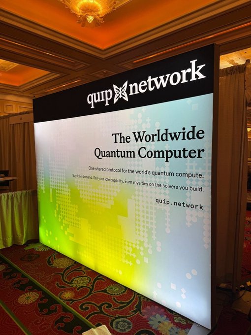 A promotional banner for Quip Network displayed at an event, reading 'The Worldwide Quantum Computer - One shared protocol for the world's quantum compute. Buy X on demand. Sell your idle capacity. Earn royalties on the solvers you build.'