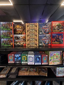 A retail display shelf stocked with various Magic: The Gathering 'PACK' boxes and other trading card game products.