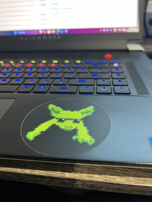 An Alienware laptop with a colorful RGB backlit keyboard and a green pixel-art alien logo sticker on the palm rest area.