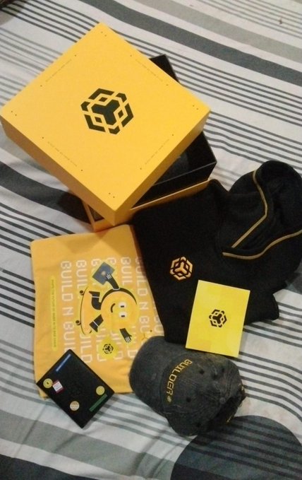 The image features a bed with various items, including a yellow box containing merchandise from BNB Chain, a black hat, a black wallet, and a black baseball cap. The items are arranged on the bed, creating an interesting display of products related to the BNB Chain brand.