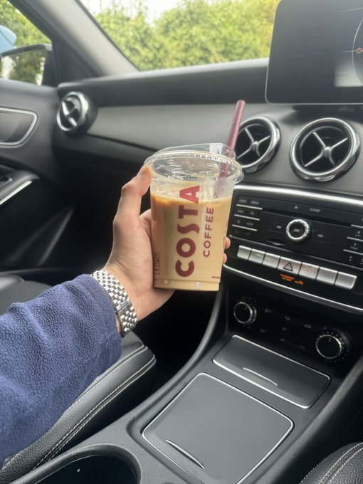 A hand holding a clear Costa Coffee cup with a pink straw sits inside a Mercedes-Benz car, featuring the dashboard with climate controls and a prominent "COSTA COFFEE" logo on the cup. The image pairs with the post "Gm family drop SOL wallets" as a casual morning coffee moment, possibly hinting at an upcoming Solana (SOL) crypto wallet announcement or launch.