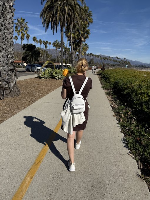 A blonde woman wearing a brown dress and a small white backpack walks down a palm-tree-lined path, captured from behind in a coastal California setting with mountains in the distance.

The image perfectly captures the "Shot from BEHIND" challenge by framing the subject entirely from the rear as she strolls down the sunlit walkway.