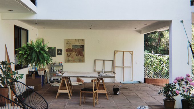 An artist's outdoor patio studio features a wooden workbench, canvases, a large fern, and potted flowers in a covered courtyard.