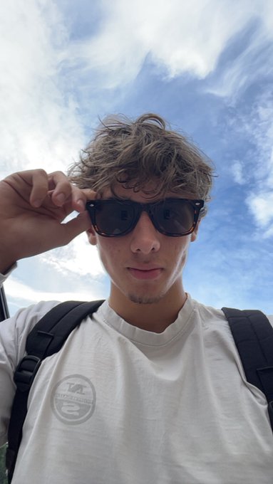 The image features a young man wearing sunglasses, a white t-shirt with a logo, and carrying a backpack. The photo appears to be taken outdoors, possibly at an early morning or late afternoon time of day when there are fewer people around. The background is filled with clouds, suggesting that the weather might be overcast. The image also contains text that reads "Good morning moggers This is the most votes i got in my mog battles so far Yall are absolute legends".