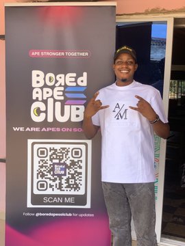 A person poses in front of a 'BORED APE CLUB' banner with the tagline 'WE ARE APES ON GO' and a QR code, wearing a white t-shirt with an 'A/M' logo.