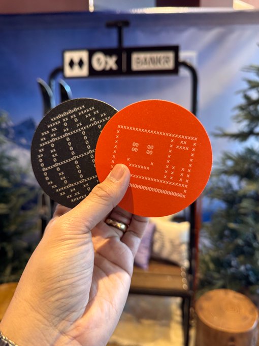 A hand holding two circular stickers—one black and one orange—both featuring ASCII art designs, with '0x' and 'DANGER' signage visible in the background.