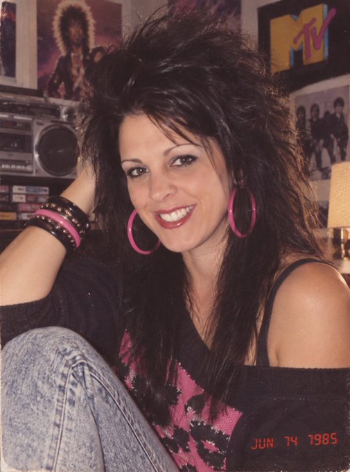 A photo dated JUN 14 1985 showing a smiling young woman with voluminous dark 80s-style hair, large pink hoop earrings, and a pink and black top, posed in front of MTV posters and a boombox.