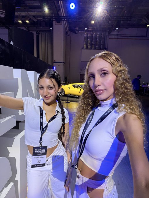 Two young women pose together at what appears to be a Singapore GP event, wearing white outfits and lanyards with one displaying a 'Giselle Jubine' credential, with a yellow sports car visible in the background.