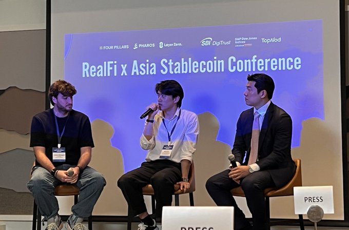 Three men seated on stage at the 'RealFi x Asia Stablecoin Conference,' sponsored by Four Pillars, Pharos, LayerZero, SBI DigiTrust, Gulf Dow Jones, and TopMod, with one panelist speaking into a microphone during a press event.