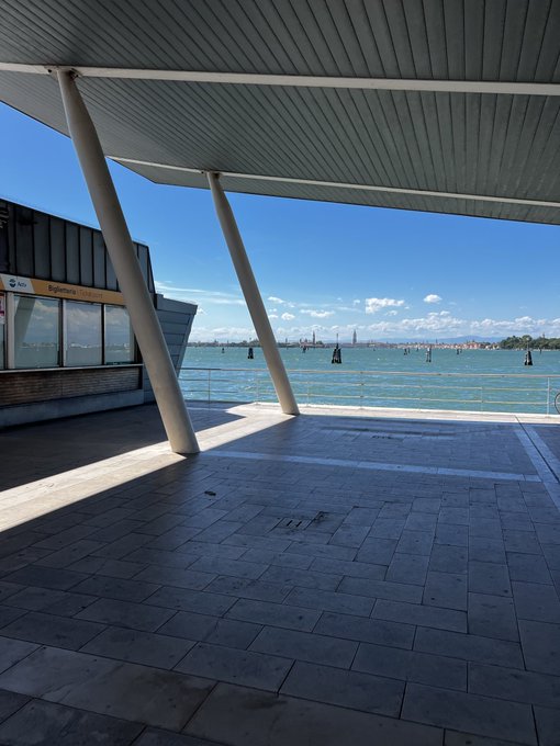 A sunny outdoor view from a modern covered waterfront terminal or pier, looking out over blue water toward a distant city skyline.
