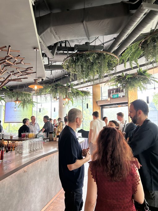 People gather at a modern, industrial-style bar or cafe with exposed ceilings and hanging plants, standing near a counter lined with glassware and bottles.