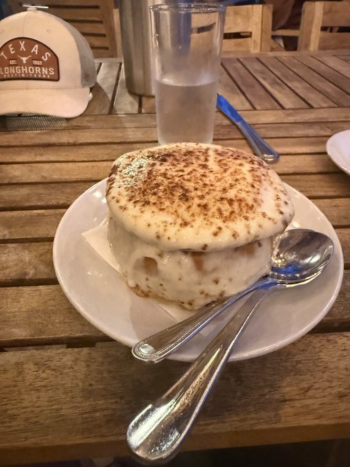 A bowl of French onion soup topped with a browned, melted cheese crust sits on a white plate at a Texas Longhorns table, paired with a glass of water and silverware. The hearty dish complements the post's promise of warm, comforting relief after a rugged day.