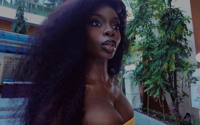 A young Black woman with voluminous hair and dark makeup poses in front of lush greenery. A sign with a red circle and line, indicating "No Smoking," is visible on the right.
