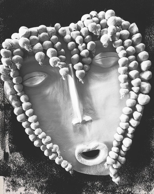 A black and white surreal sculpture of a face covered in small round objects resembling garlic bulbs, with elongated features and closed eyes.