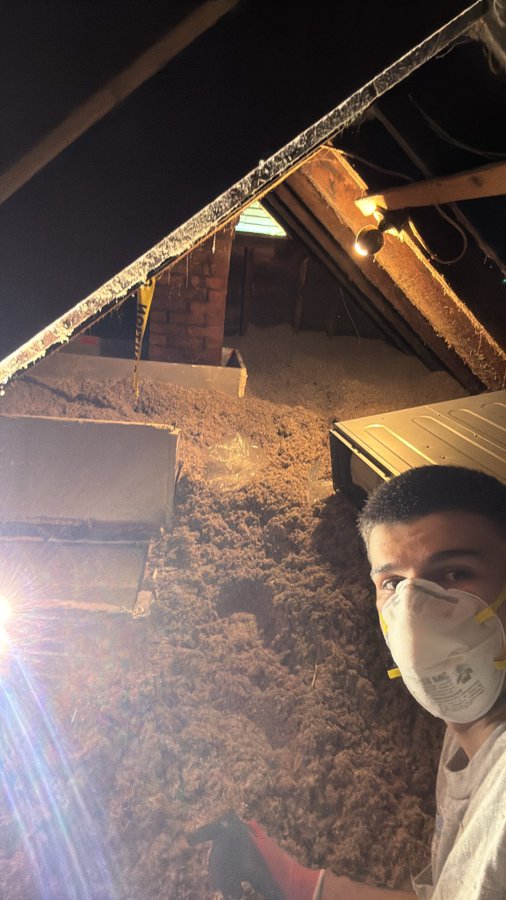 A selfie in an attic or shed at night, showing a man wearing a 3M respirator mask surrounded by blown-in cellulose or fiberglass insulation, with a 3M dust mask visible bearing the "3M" logo. The image humorously ties to "$DRB is for the people" by suggesting a grueling, sweaty insulation installation job—likely illustrating the meme's theme of hard, unglamorous work being done "for the people."
