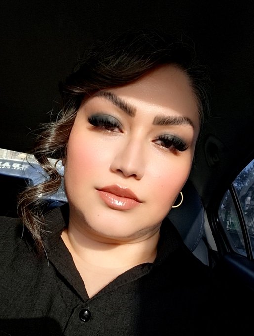 A young woman with dramatic smoky eye makeup, bold brows, and glossy lips takes a selfie inside a car, with natural light illuminating her face against the dark interior. Her polished, high-fashion look perfectly fits the caption's humorous tone of "the woman you buy NFTs from," giving off a stylish, influencer-like vibe.