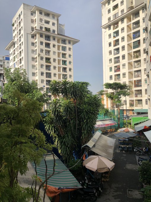 A view of tall white apartment buildings surrounding a courtyard filled with tropical trees, green awnings, umbrellas, and parked motorbikes.