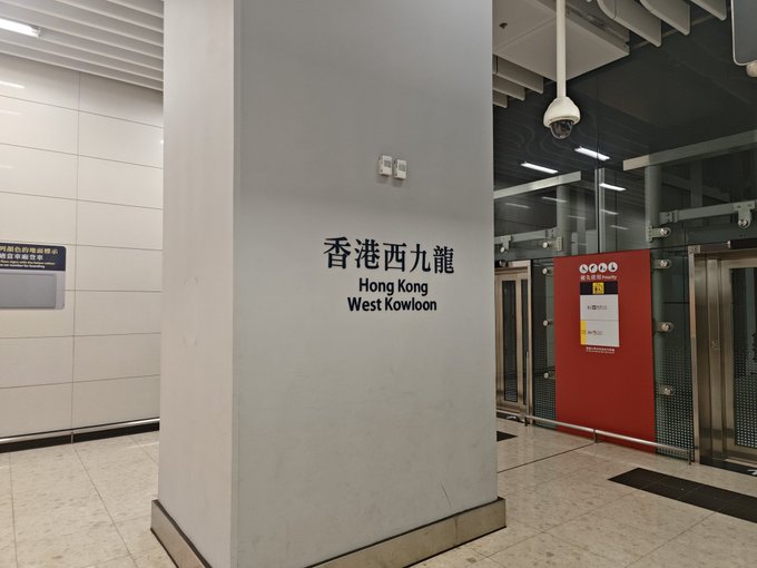 The image shows a pillar at the Hong Kong West Kowloon station with the name displayed in both Chinese (香港西九龍) and English. The caption reflects the traveler's departure from Hong Kong after a productive trip opening bank accounts (HSBC, ZA Bank, etc.) and meeting friends, with this station serving as the departure point from the city.