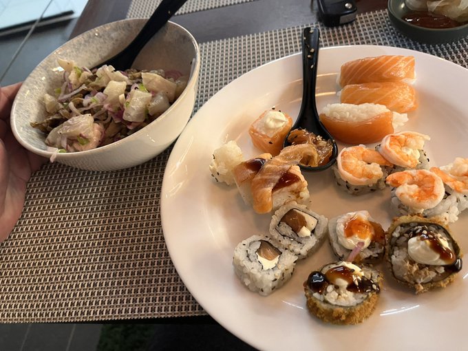 A meal consisting of a bowl of pasta salad and a plate with various sushi rolls and sashimi, accompanied by a black spoon.