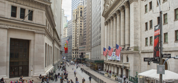 A street view looking down Wall Street in New York, with American flags hanging on the neoclassical building (Federal Hall) and pedestrians walking below.