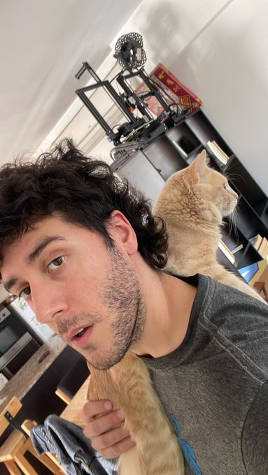 A young man with dark curly hair holds a tan cat on his shoulder in a kitchen setting, with gym equipment visible in the background.