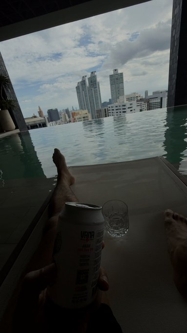 A person relaxes by an infinity pool holding a beer can, with their feet resting on the edge overlooking a city skyline.