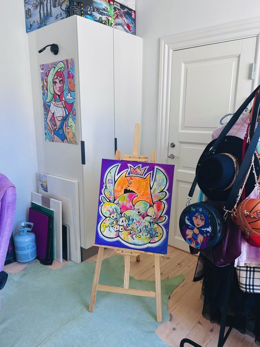 An artist's studio with a colorful, abstract pop-art painting displayed on a wooden easel, alongside other artwork, bags, and a white cabinet.