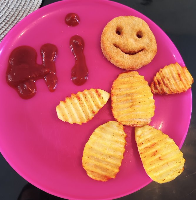 A pink plate with food art made of ketchup writing 'HI' and a smiley face formed by a breaded nugget, with crinkle-cut potato pieces arranged like a body below.
