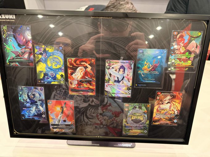 A display case showcasing trading card game cards with anime-style artwork, including characters like Leviathan Vanguard and Phoenix, with the photographer's reflection visible in the glass.