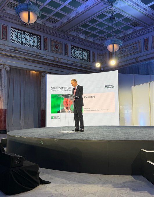 A man stands at a clear podium delivering a 'Keynote Address' on a circular stage, with a presentation screen behind him showing 'Paul Atkins' and the 'SS&C' and 'Washington Wall Street' logos.
