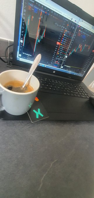A coffee cup sits beside an HP laptop displaying active trading charts, with an xPortal card (marked with a teal "X") placed in front, matching the post's mention of needing the @xPortalApp card to complete the setup. The trader is monitoring $EGLD (MultiversX) charts while enjoying their steaming coffee.