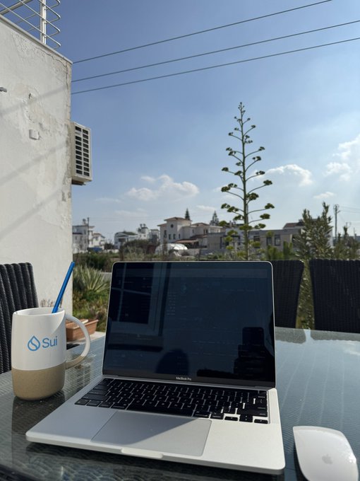 A MacBook Pro sits on an outdoor glass table next to a white and tan "Sui" branded mug, with a view of buildings, plants, and a blue sky.